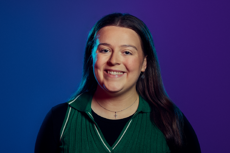 Headshot of a dark haired young female wearing a green v-neck over a black long slieved shit, standing in front of a blue to purple background